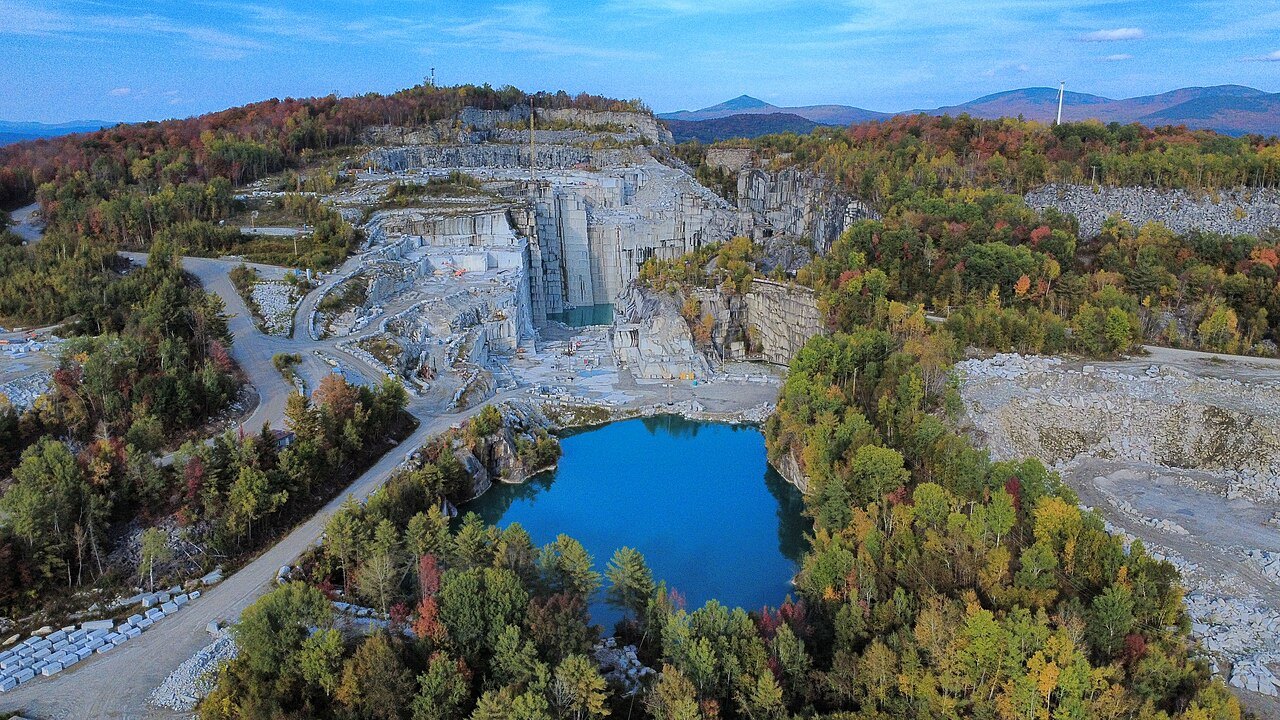 The pit of the Rock of Ages quarry, filled with blue water below white granite benches, with autumn color beyond.