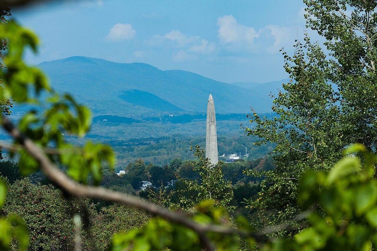 The monument seen from a wooded overlook, its pale obelisk small against blue mountains.