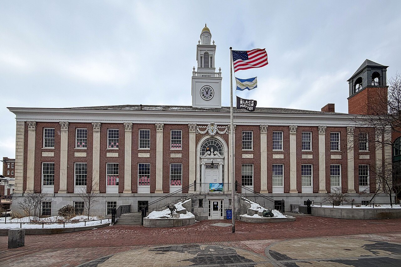 Burlington City Hall, a brick building with tall windows and a clock tower, flying the flag.