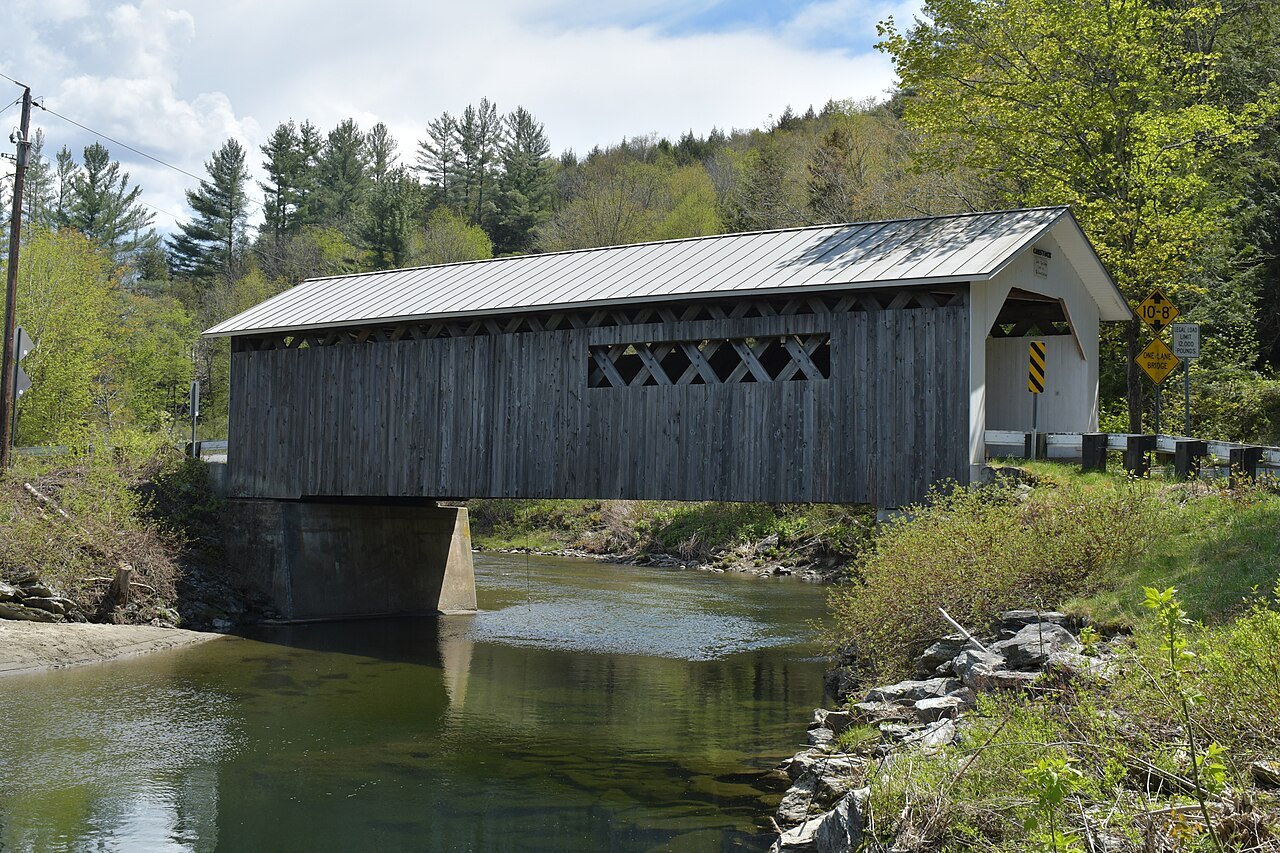 The Comstock Covered Bridge, a gray bridge with a white-trimmed roof, over a calm river.