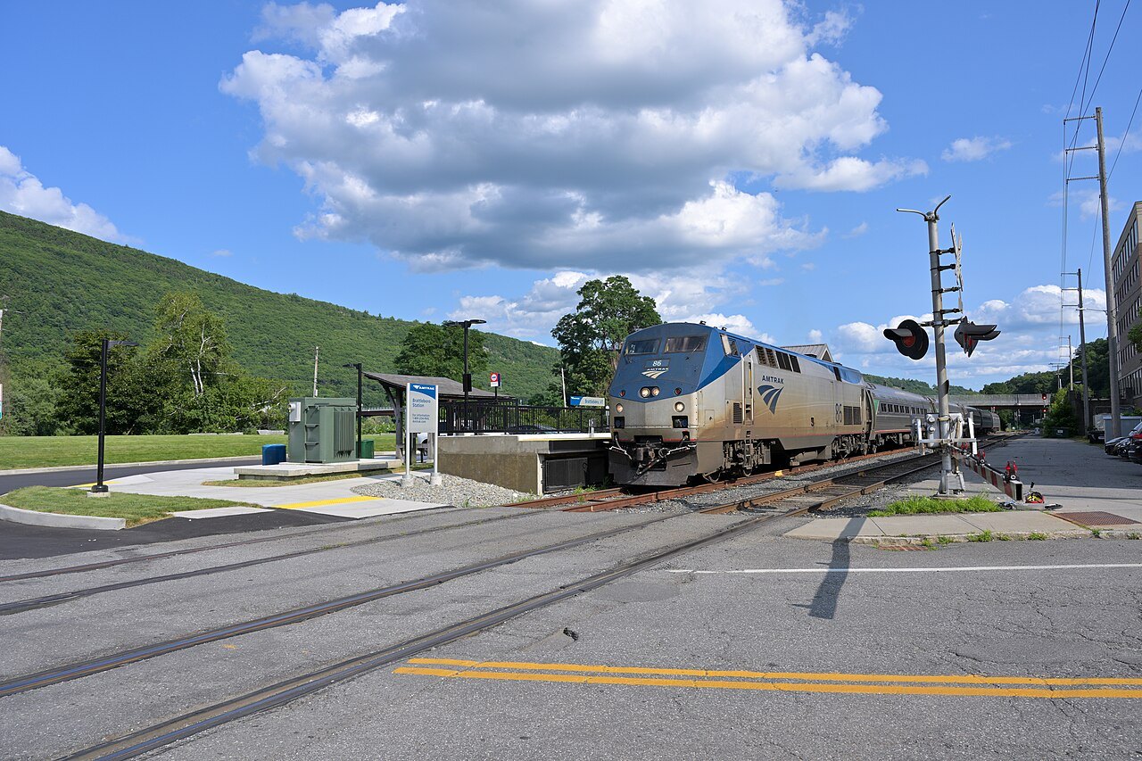 An Amtrak Vermonter train stopped at the Brattleboro platform, with green hills behind the station.