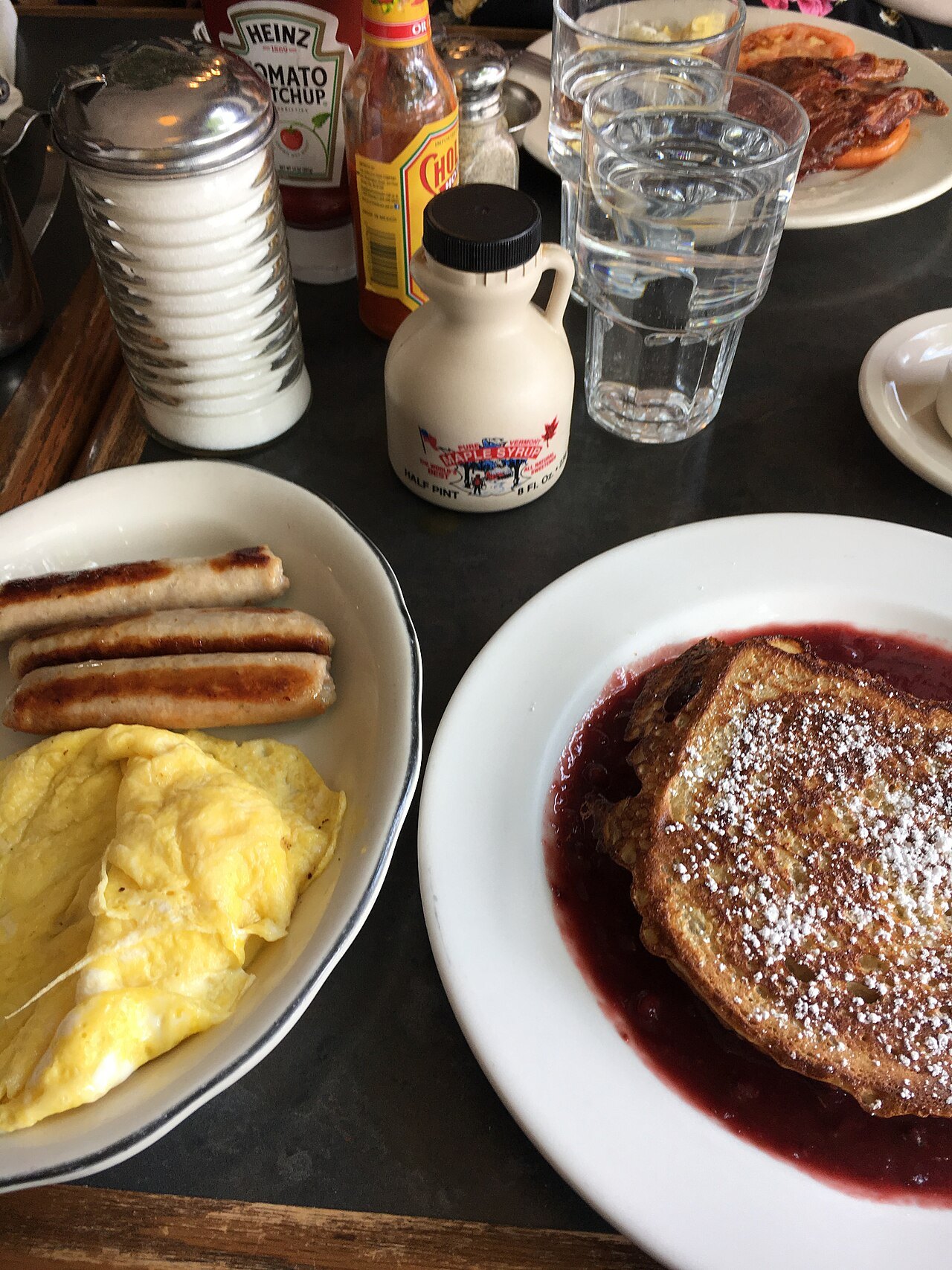 A breakfast table with French toast, scrambled eggs and a bottle of maple syrup.
