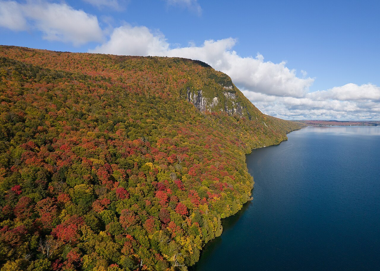 Lake Willoughby from above in October, a cliff and a slope of red and orange forest above blue water.