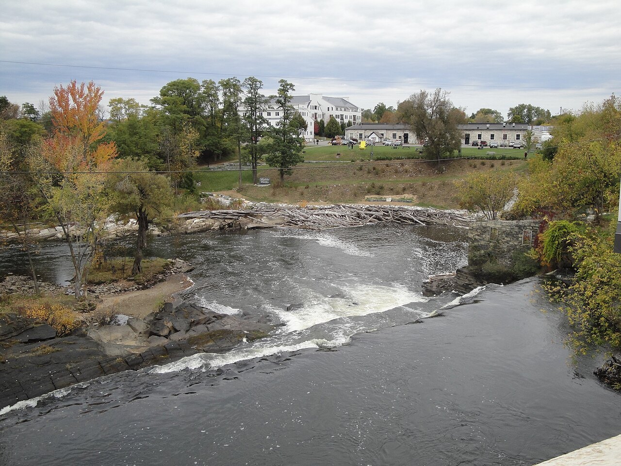 Otter Creek rushing over a ledge of rock at the falls, with a white building beyond.