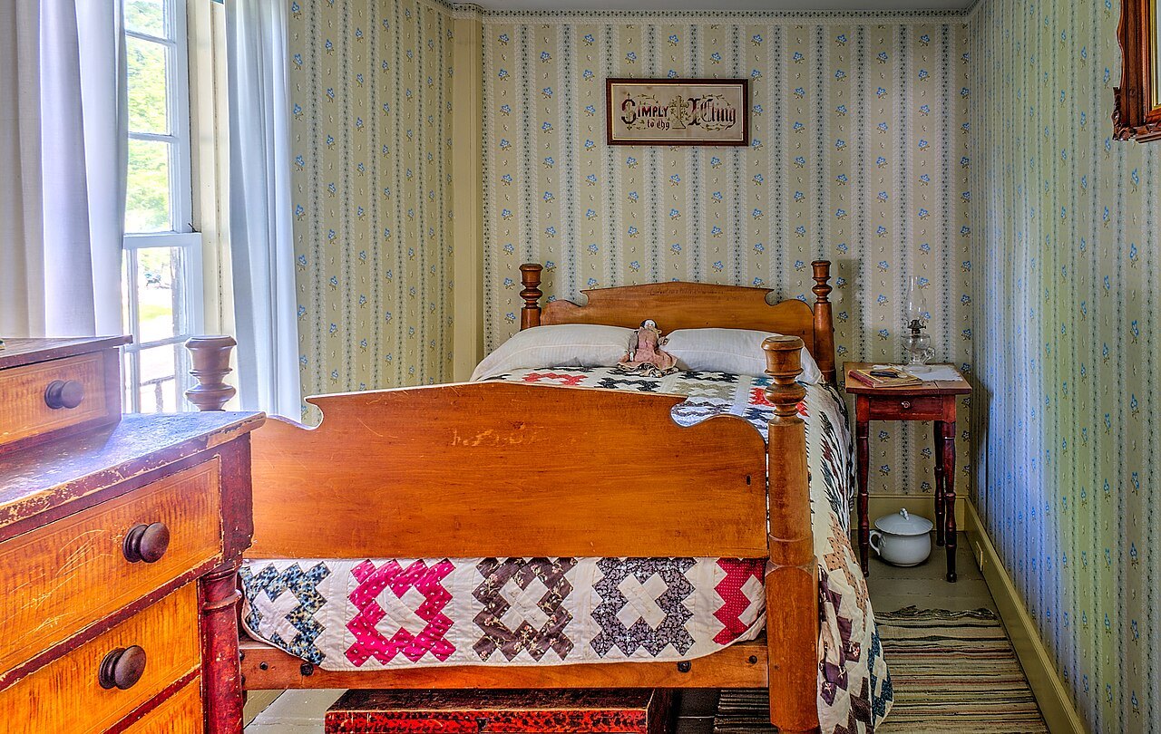 A bedroom with a red-and-white quilt, striped wallpaper and a washstand.