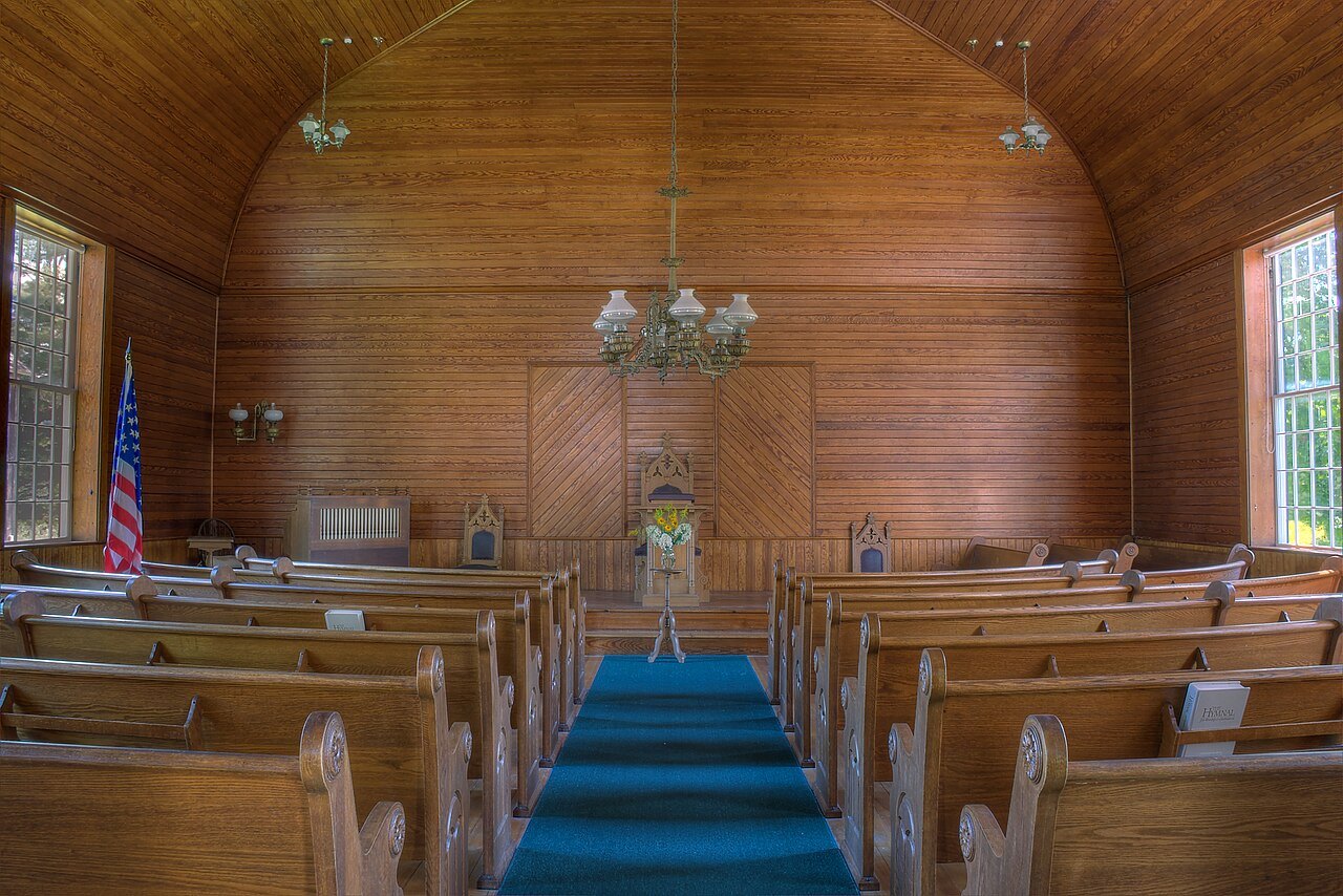 The interior of the village church, with wooden pews, a blue aisle runner and a paneled apse.