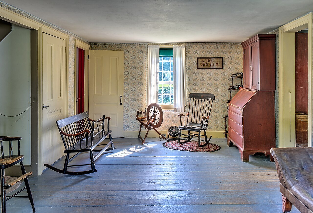 A sitting room in the homestead, with rocking chairs, a spinning wheel and a slant-front desk.