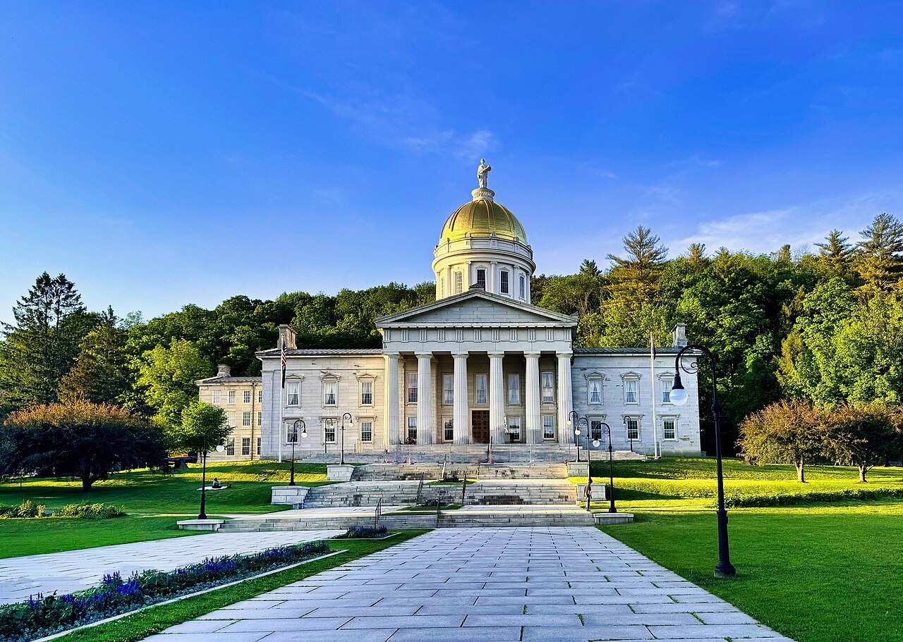 The Vermont State House from its walk, with the gold dome above flower beds and a blue sky.