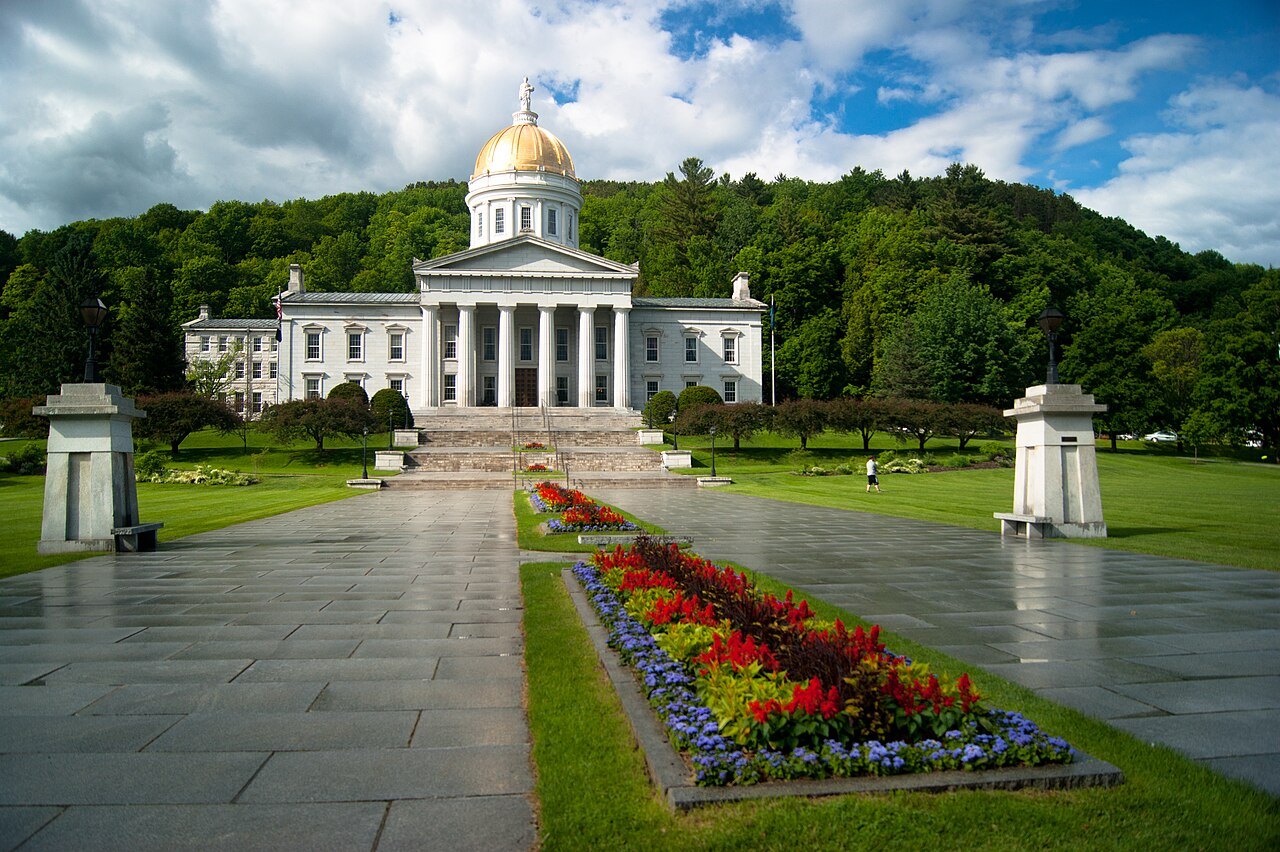 The State House beyond a wet stone walk and beds of red and yellow flowers.