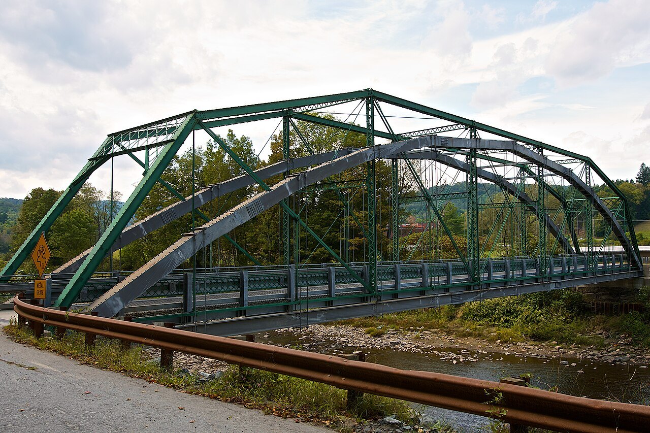 The West Woodstock Bridge, a green steel truss bridge, with a guardrail in the foreground.